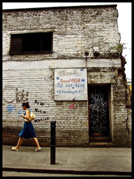 An unlikely location for a social club; a lady hurries past, unconsciously mirroring the colours of the sign - the crumbling East End.