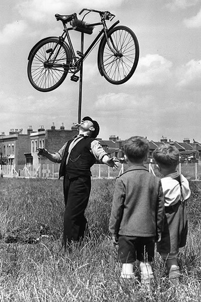 Balancing Act: 16 August 1952: In the field outside his house in Plaistow, East London, 62-year-old railway worker Mo Harris balances a bicycle on top of a broom on his chin, watched by a couple of incredulous youngsters. Harris is known as "the chin-up boy of Stratford" (Photo by William Vanderson/Fox Photos/Getty Images) 