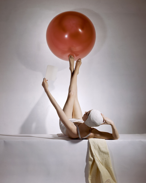 Reclining model in white swimsuit and bathing cap, balancing large red ball on feet