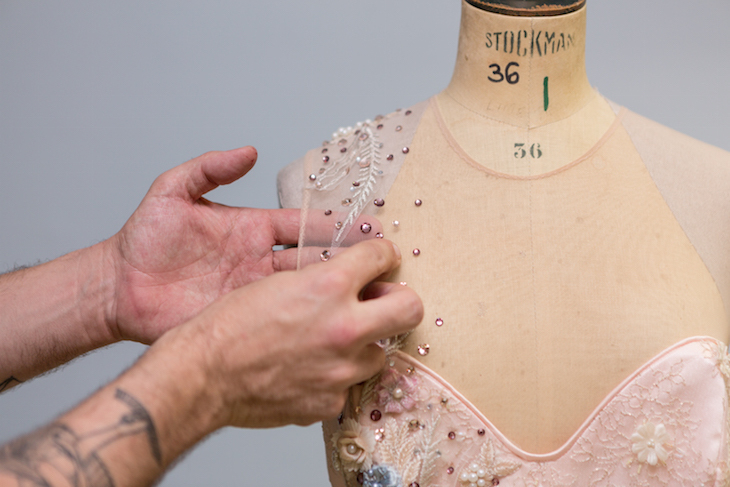 Close-up shot of a hand sewing sequins onto the shoulder of a pink dress on a mannequin, in National Theatre's wardrobe department