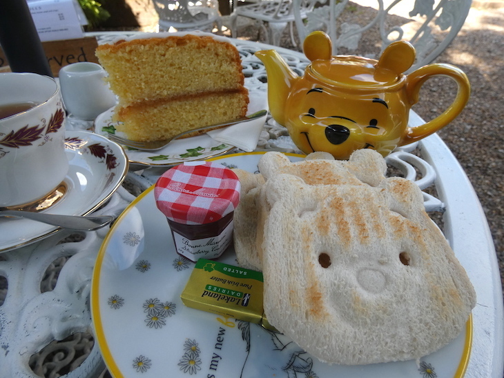 A plate with toast in the shape of Winnie the Pooh's face, alongside a slice of Victoria sponge cake, and a teapot shaped like Winnie the Pooh's head.