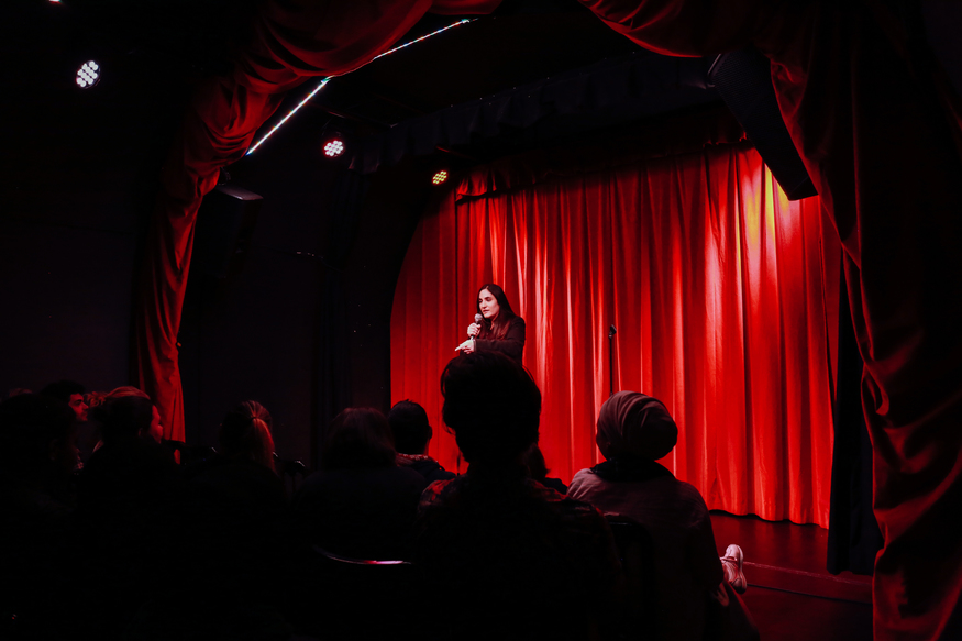 jenan performs in front of a red curtain at a Weapons of Mass Hilarity night
