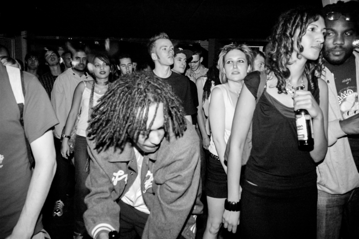 A dreadlocked man in the throes of DJing with a party crowd behind him
