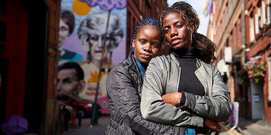 two young woman embrace on a street while looking defiantly down the lens