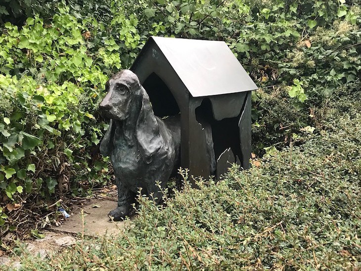 A dog in a kennel, both made of bronze. They lurk in some bushes.