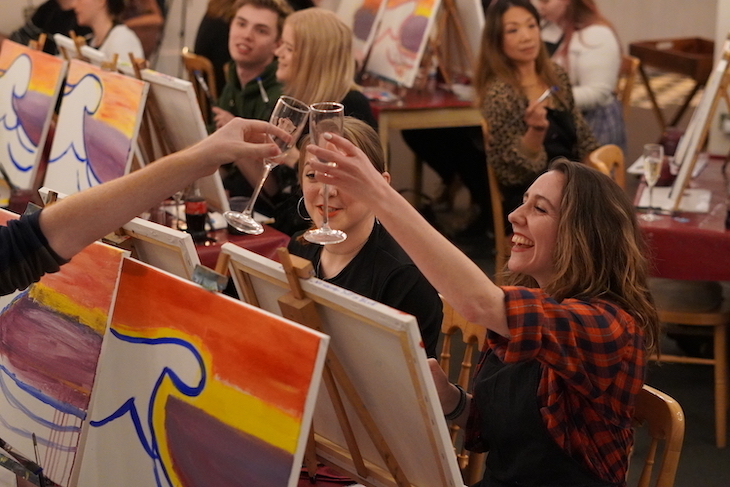 A group of smiling painters in front of their canvases. The nearest two are toasting each other with Prosecco glasses