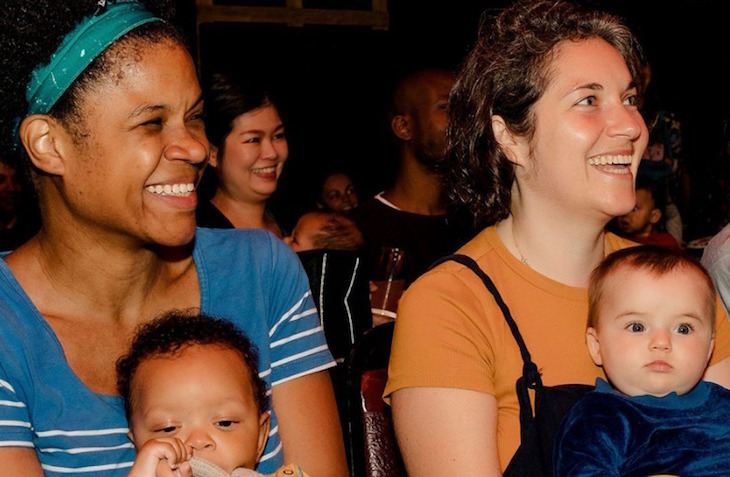 Two women sitting next to each other, looking at a stage and laughing. They are each holding a baby.