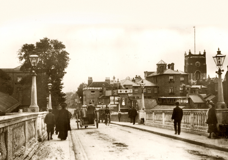 A very old black and white photograph showing people crossing a bridge towards a town with a prominent church