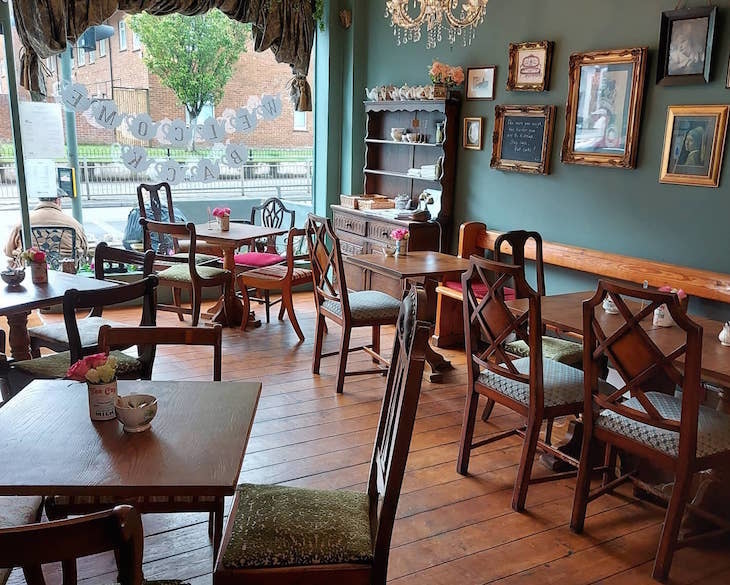 Interior of Earl Grey Tea Rooms; Teal coloured walls, wooden chairs and tables, wood floors. Framed pictures on the wall and vases of flowers on some tables. The window at the end looks out onto the main road.