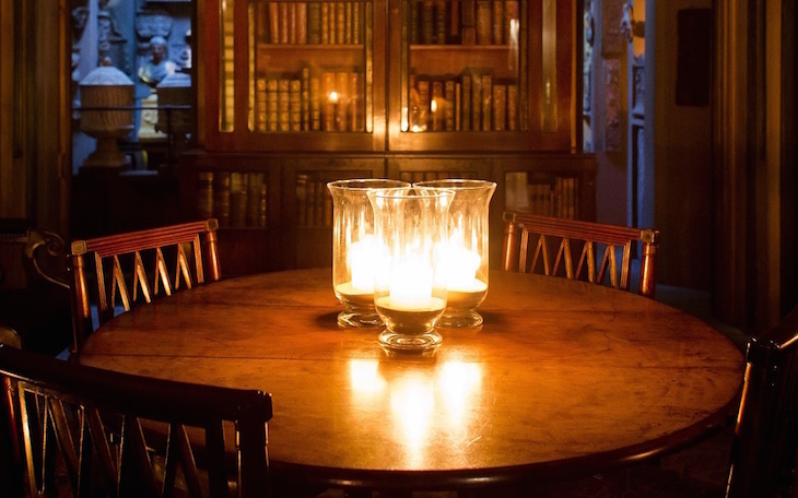 Three candles in hurricane vases on a wooden table in an otherwise dark room, with a bookcase behind.