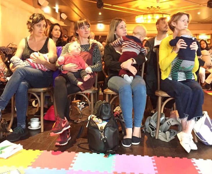 Four women sitting in the front row of an audience, each holding a baby, with a soft play mat on the floor in front of them