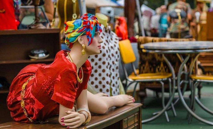 A mannequin in a red dress and multicoloured headscarf, lying on a wooden chest of drawers, with other mannequins and items of furniture in the background