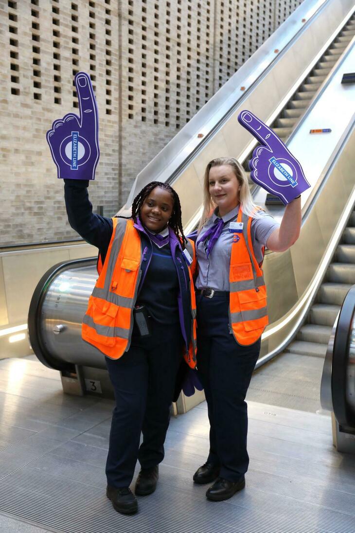 Two Elizabeth line staff in high vis wave giant purple foam hands