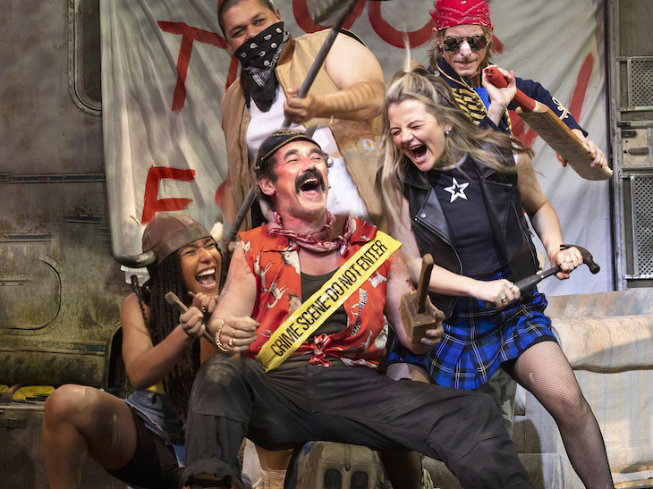 5 people, eccentrically dressed, appear to bellow and laugh on stage. The foremost wears a police tape sash saying crime scene do not enter