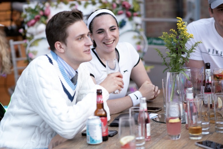 A young man and woman wearing full tennis gear enjoy a drink