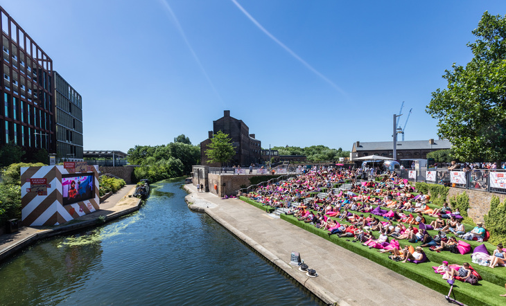 People sit in the sun on the ghat steps overlooking a canal - on the other side of the water is a huge screen