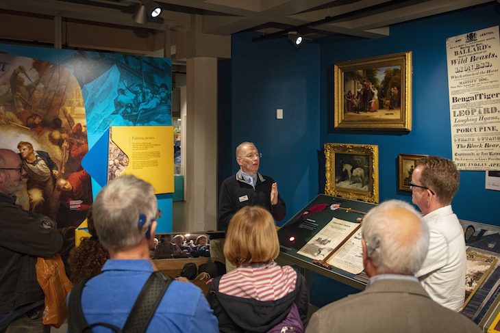 A group of museum-goers look on as a guide tells them about nearby paintings on a blue wall, using sign language