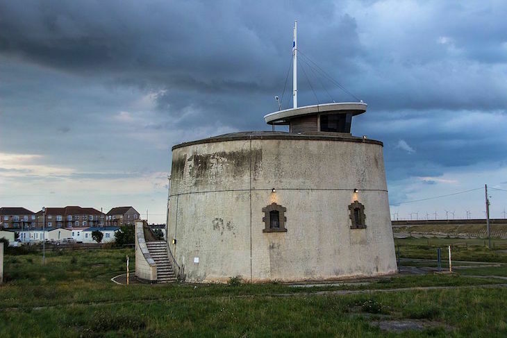 Jaywick Martello Tower: A beige concrete martello tower, as wide as it it tall, with a flight of stairs going up one side, and a viewing gallery with mast on the roof.