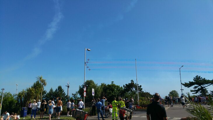 5 planes are seen flying in formation in the sky. They have blue and red fumes coming from their tails. A crowd are looking up at the sjy to see them. 