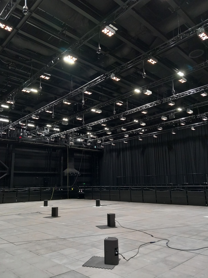 A number of speakers are placed on one of the experimental stages. The hundreds of specialist lamps are attached to lighting rigs high up on the ceiling. The walls are black and the floor is grey concrete. 