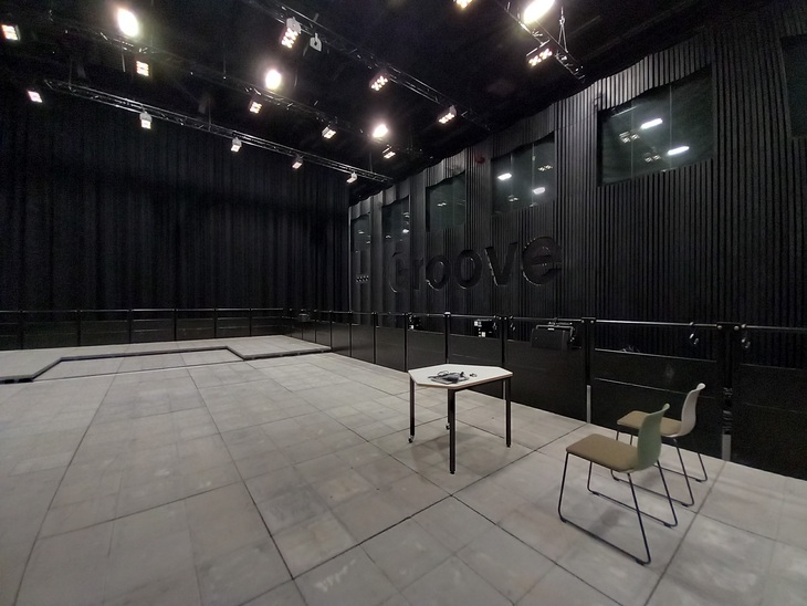 A black room with a concrete floor. 2 chairs and a table are placed on the concrete floor to the right of the image. The word "GROOVE" is embossed on the wall in the background