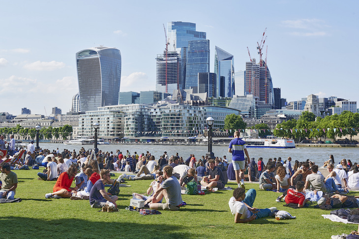 People sitting in the sunshine alongside the Thames at Summer by the River, with the Square Mile skyscrapers in the background