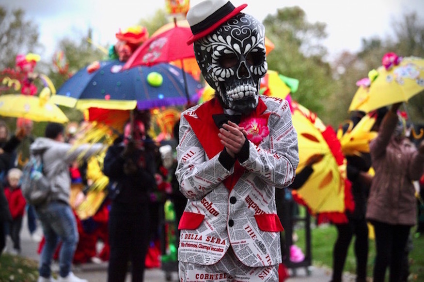 A person dressed in a newspaper print suit, with a black and white Day of the Dead style skull mask. People in the background hold colourful umbrellas, as if at a festival.