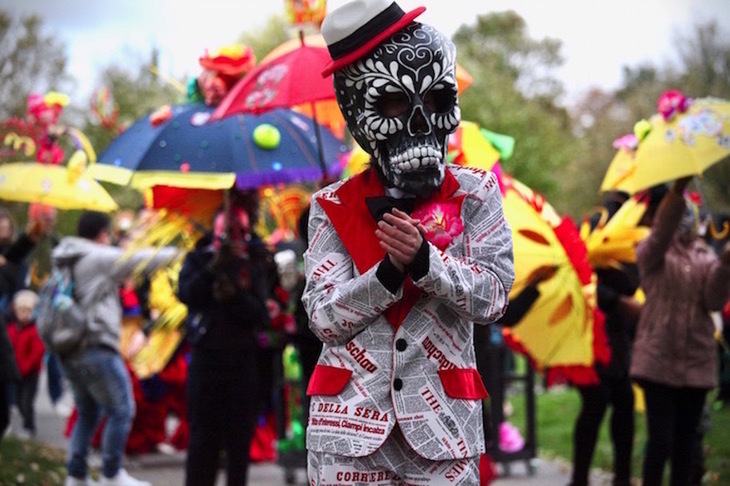 A person in a newspaper suit wears a huge black day of the dead mask