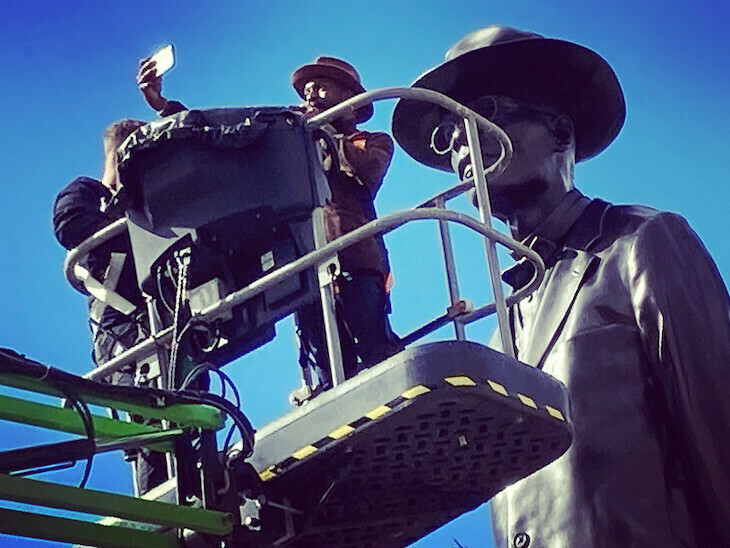 A statue of a man in a hat, with the artist in a similar hat taking a selfie from a cherry picker
