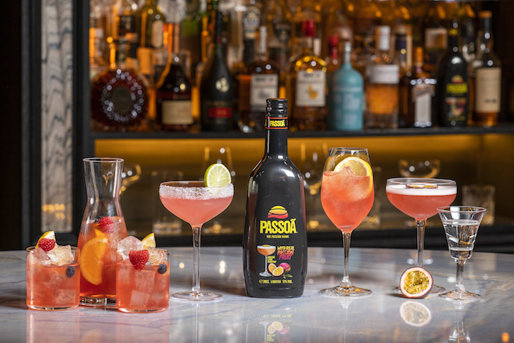 A selection of cocktails in various different glasses lined up a grey marble top bar, with a black bottle of Passoã liquer in the centre.