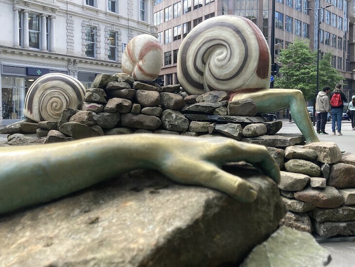 A metallic hand clutches a rock in foreground, with several snail-human hybrids in the background. 