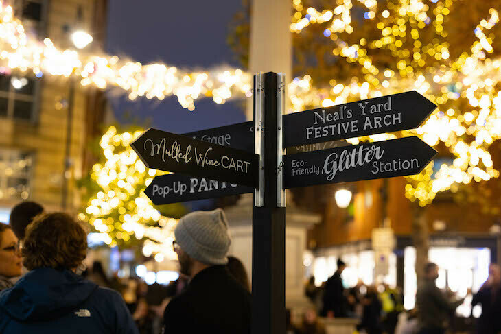 People in winter clothes gathered underneath fairy lights, and a signpost pointing in different directions to 'Neals Yard Festive Arch', 'Glitter Station', 'Mulled Wine Cart' and other festive activities