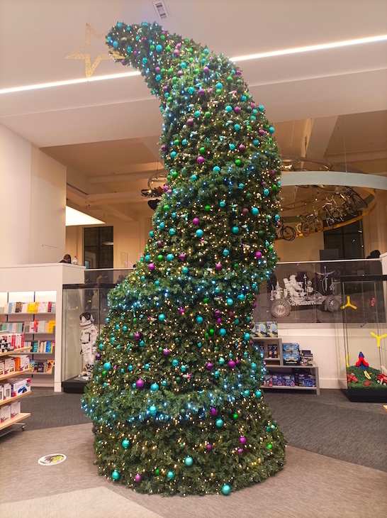A tall thin Christmas tree bending in as couple of places. It's decorated with blue, green and purple baubles.