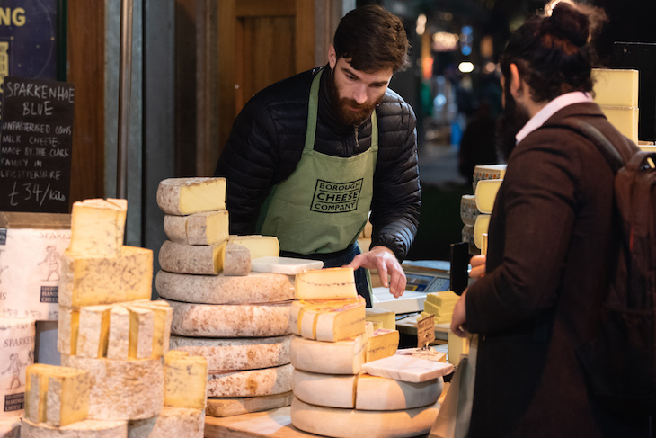 A cheesemonger attending a stall piled high with cheese, as a customer browses