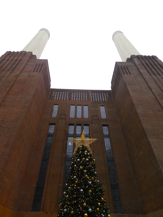 A tree covered in red, silver and gold baubles and topped with a gold star,  in front of the chimneys of Battersea Power Station