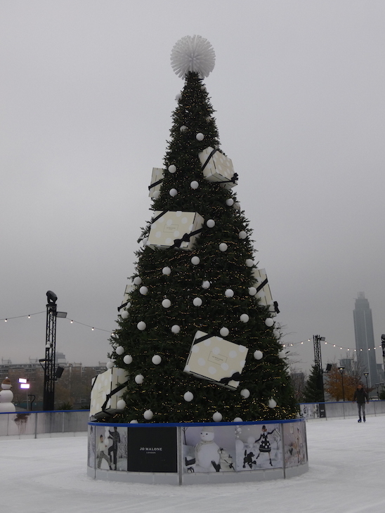 A tree in the middle of an ice rink in daylight. It's covered in white bauble and large white Jo Malone gift boxes, with a large white pom pom on top