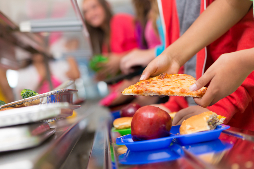 A school canteen, showing hands grabbing out for food including a pizza slice and apple