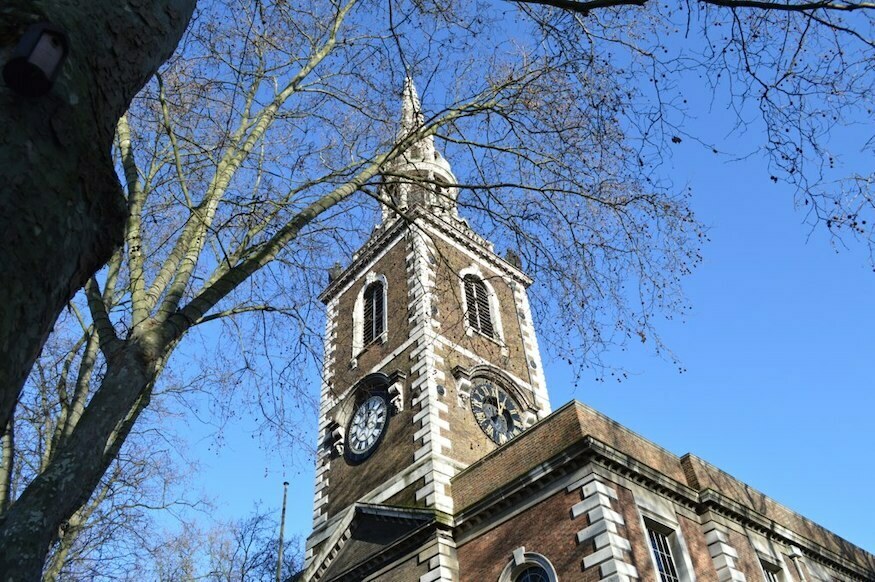 The brown and white steeple of a church rises into a clear blue sky. A defoliated tree is in the foreground