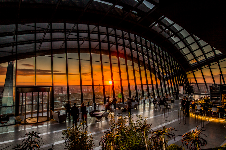 The floor-to-ceiling glass windows of the Sky Garden, looking out to an orange sky as the sun sets over London.