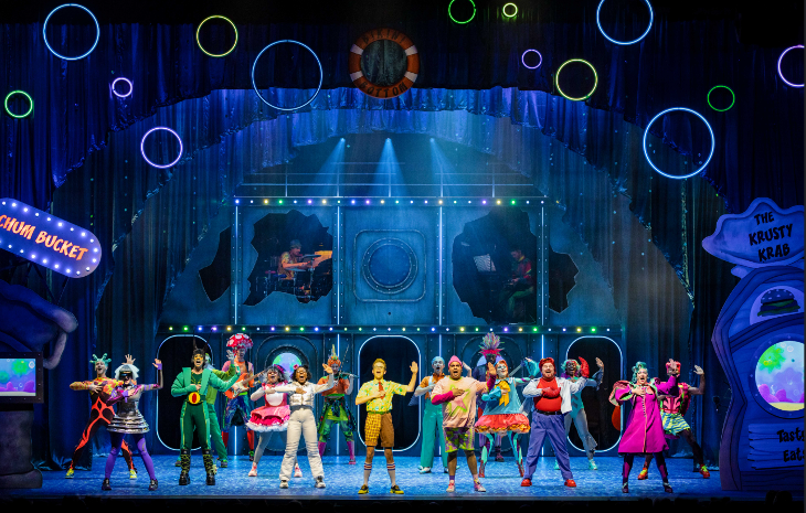 The cast of Spongebob The Musical mid-performance, lined up along the front of the stage, each raising one hand in the air.