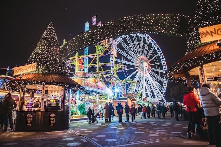 A bridge of fairy lights across a path, with wooden market huts on either side, a giant Christmas tree in the foreground and the big observation wheel in the background, all lit up at night