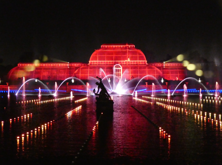 Kew's Palm House illuminated red at night, with fountains dancing on the lake