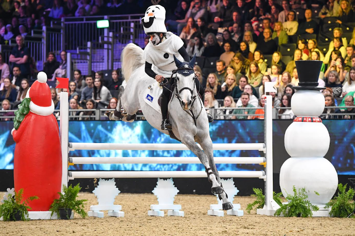 A rider wearing a snowman hat riding a horse over a jump, with Santa and snowman decorations on either side