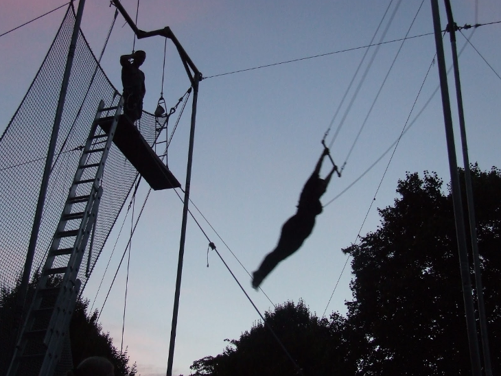 A silhouetted photo of someone mid-air swinging on a trapeze bar, with trees in the background