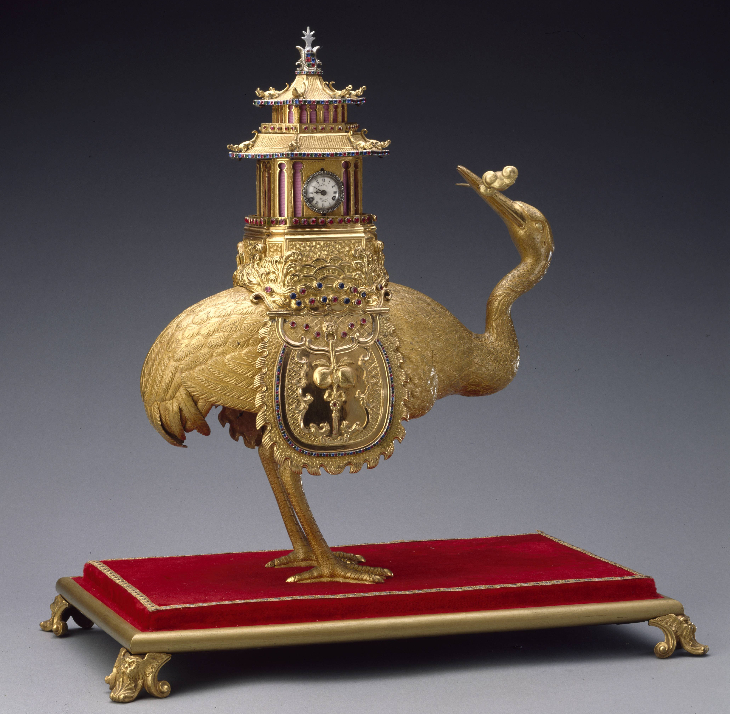 A golden clock on the back of a golden ostrich, displayed on a red carpeted plinth