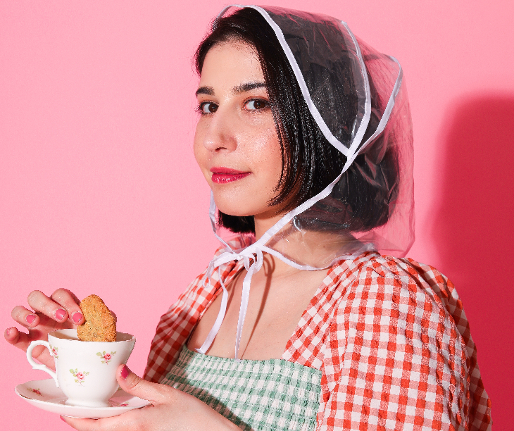 Priya Hall in a gingham dress, wearing a plastic rain hood and holding a cup of tea and a biscuit