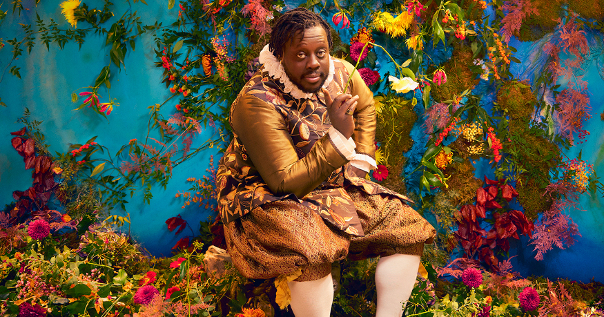 An actor in period costume in front of a wall of flowers