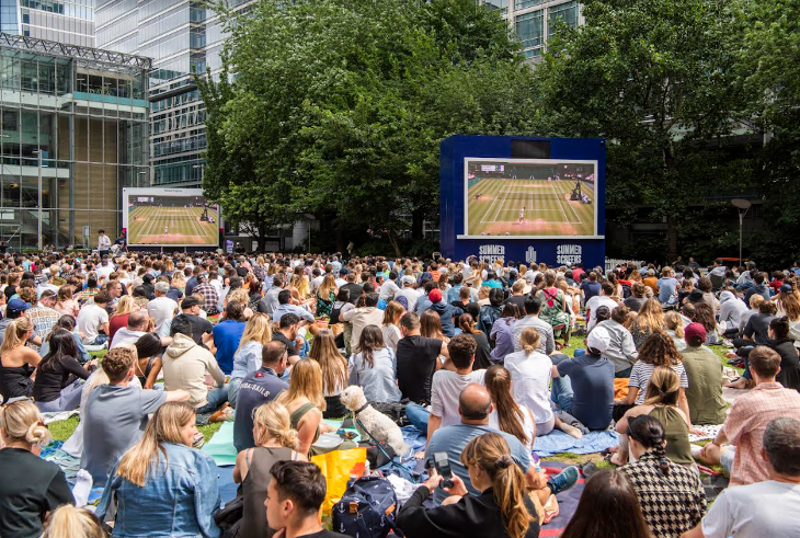 People sitting on the grass and on picnic blankets watching Wimbledon on a large screen in Canary Wharf