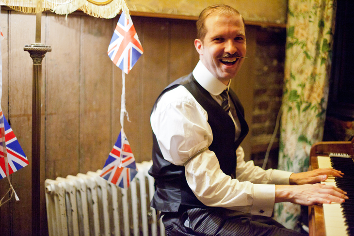 A man in a waistcoat plays piano with union flags behind