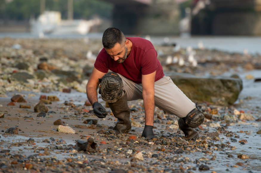  Mudlarker Alessio Checconi with knee pads and gloves crouches on a rocky shoreline